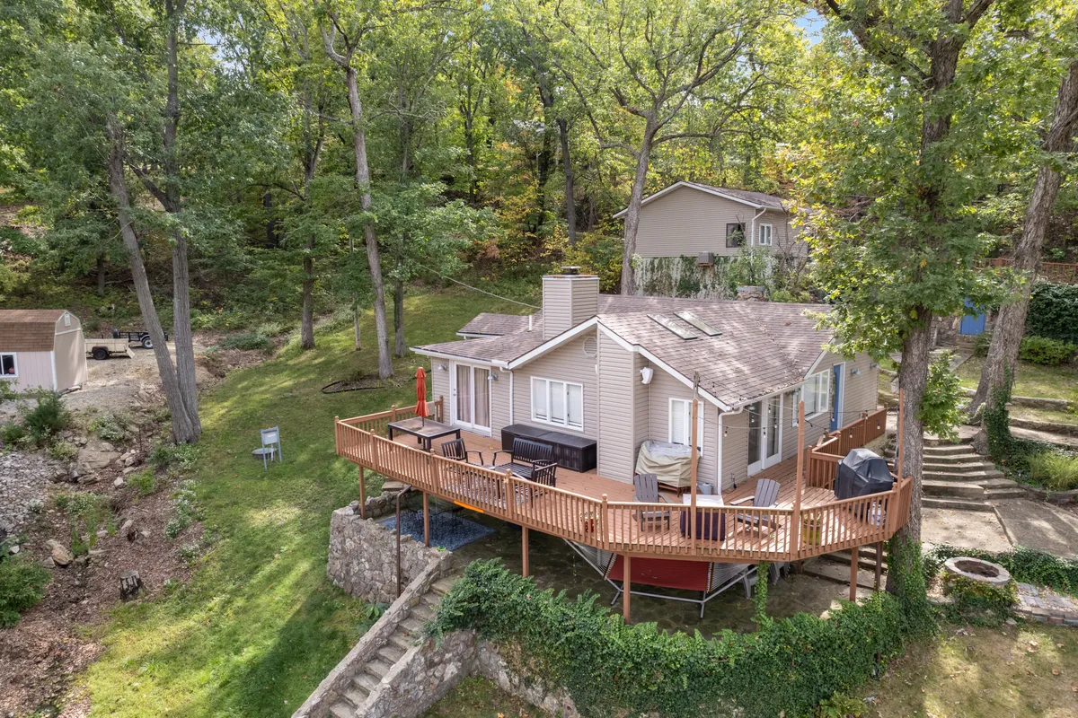 A spacious outdoor wooden deck surrounded by trees with a comfortable seating area and a sturdy stone wall. The setting is ideal for gatherings, barbecues, or relaxing after a day out, with the sunset filtering through the trees and an inviting, communal atmosphere.