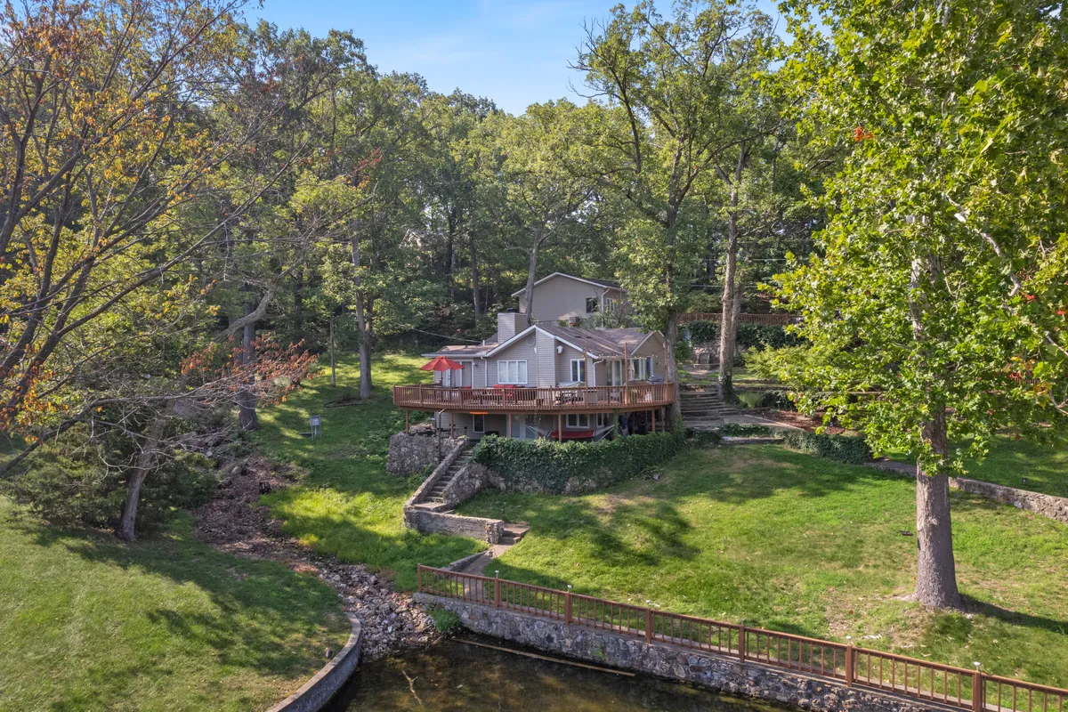 A scenic view of the house with its deck and umbrella, stone steps, plentiful trees, and immediate access to the waterfront area. The image captures both the home base and the surrounding nature, suggesting proximity to trails and the lake for all types of outdoor fun.
