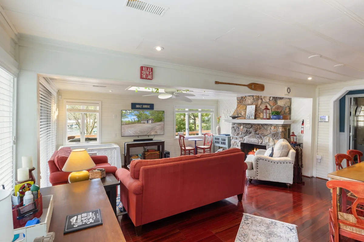 A spacious interior with comfortable red sofas, a substantial stone fireplace, and expansive windows that reveal scenic views outside. Decorative touches like a paddle on the wall enhance the vintage lakeside theme, while the layout promotes conversation, games, and family time.