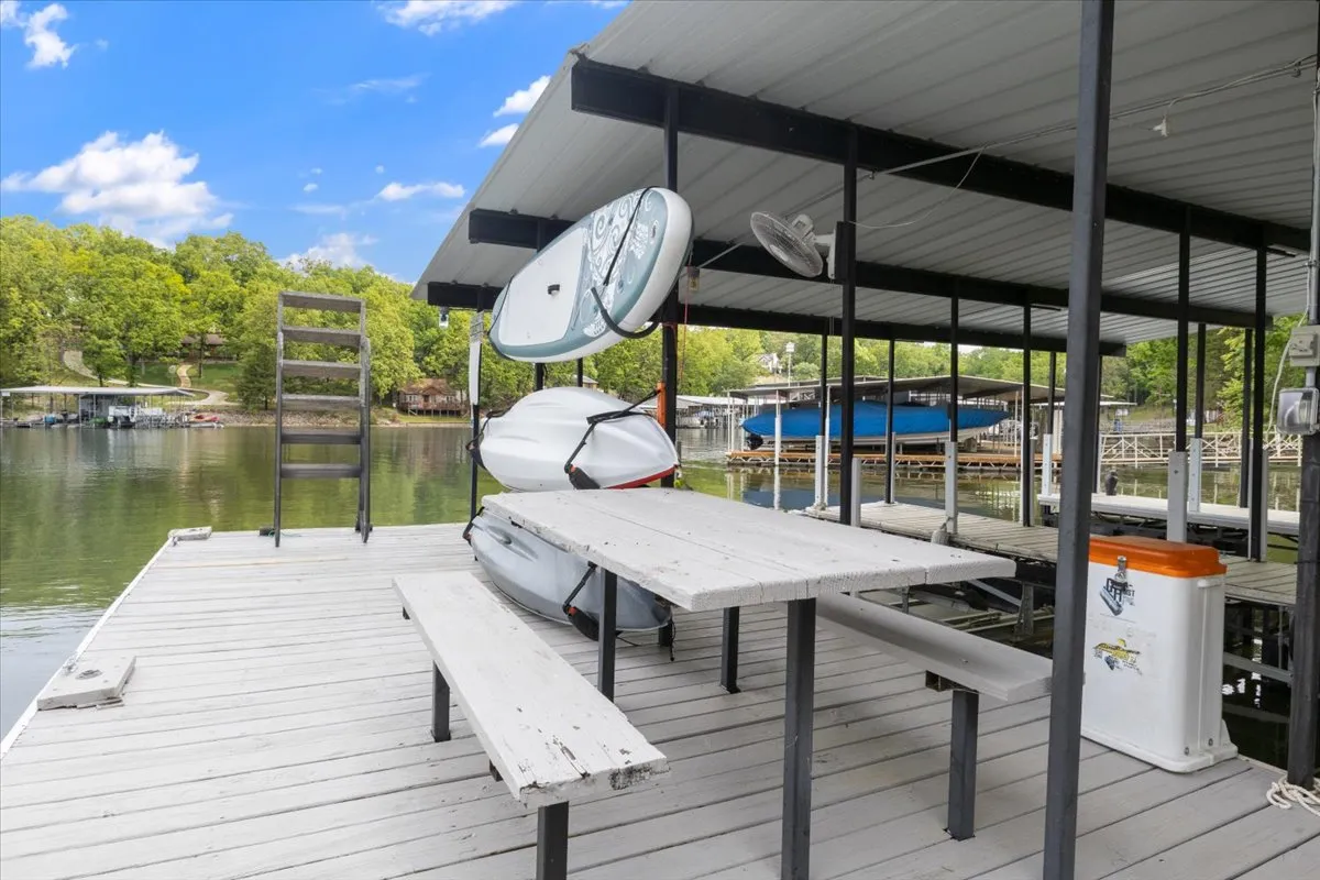 A tranquil scene of a private dock extending over calm water, featuring a kayak pulled up along the side, dock seating, and lush greenery along the shoreline. The setting suggests ease of access for water activities and relaxation on the dock itself.