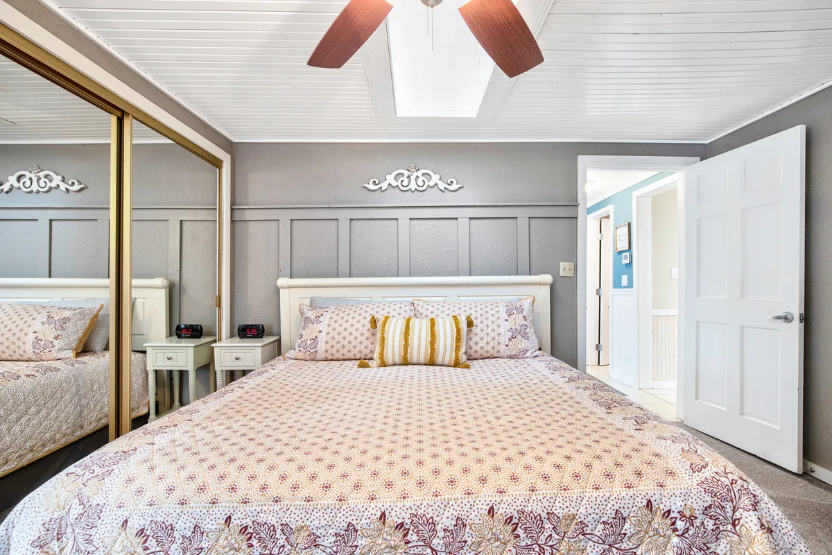 A comfortable bedroom featuring a bed with plush bedding, a mirrored closet, a ceiling fan, and modern wall decor. Ample natural light fills the room, contributing to a bright, relaxing environment.