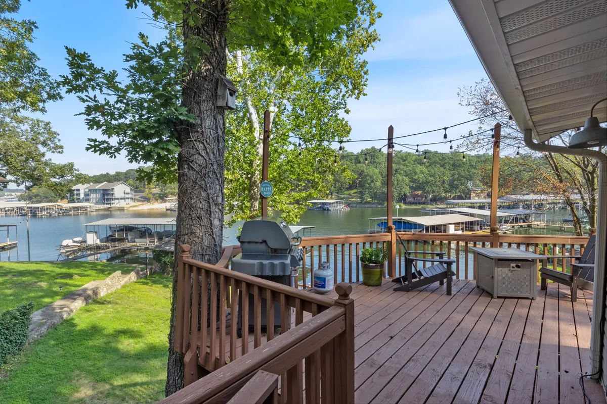 A spacious wooden deck overlooks the sparkling lake, featuring a built-in grill, comfortable seating area, and ample space for gatherings. The deck is positioned to capture beautiful water views and is surrounded by lush greenery, making it ideal for indoor-outdoor living.
