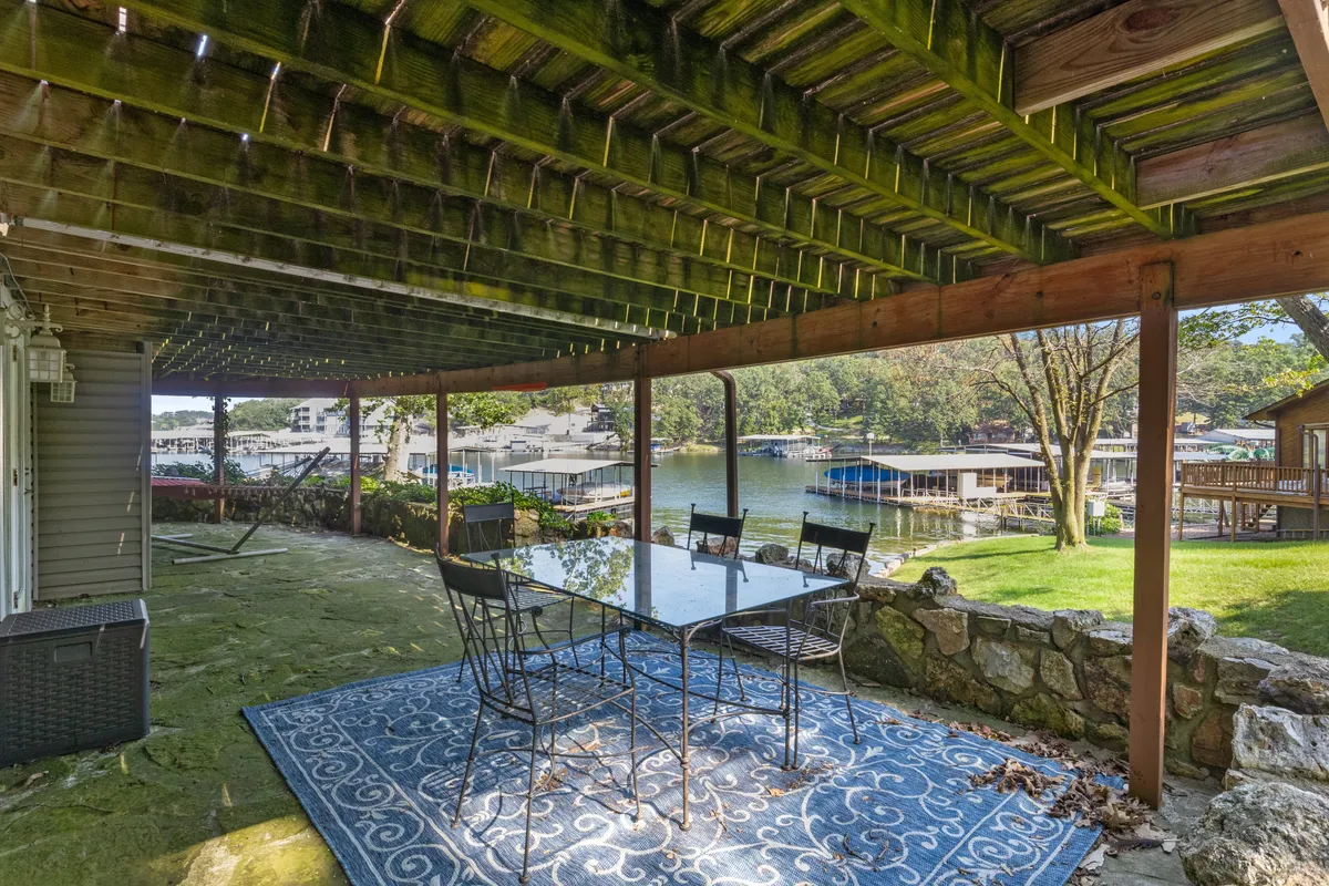A stone patio with a glass dining table and black chairs overlooks the lake, framed by mature trees. The scene captures the impressive lake views visible from the home and the seamless connection between indoor comfort and outdoor beauty.